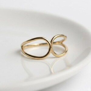 May include: A gold ring featuring an open, asymmetrical design composed of two distinct, smooth, rounded loops of varying sizes that are joined together at the centre, creating a continuous, polished metal band with a reflective surface.