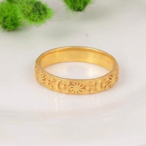 May include: A gold-toned band ring featuring an embossed pattern of suns, crescent moons, and small stars encircling the entire outer surface. The ring has a smooth, polished finish and a consistent width throughout its circular design.
