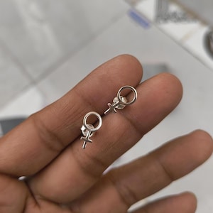 Puede incluir: Un par de pendientes de plata con el símbolo femenino. Los pendientes son pequeños, con una parte superior circular y una cruz debajo. Se muestran sobre una superficie clara.