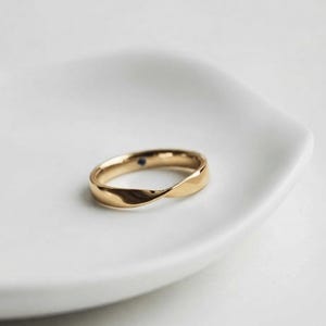 Op de afbeelding: Een gepolijste gouden ring met een subtiel gedraaid ontwerp dat een doorlopende vloeiende vorm creëert. Het metalen oppervlak is glad en reflecterend en een kleine donkerblauwe edelsteen is aan de binnenzijde van de ring gezet.