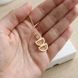 Op de afbeelding: Een goudkleurige ketting met een hanger bestaande uit twee op elkaar gestapelde halve cirkels. De hanger hangt aan een fijne, dunne goudkleurige ketting. Het ontwerp bestaat uit een bovenste halve cirkel die naar boven wijst en een onderste halve cirkel die naar beneden wijst, wat een symmetrisch, open geometrisch motief vormt.