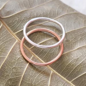May include: Two thin, circular rings stacked together. One ring is a polished silver-tone metal, and the other ring is a polished rose gold-tone metal. Both rings feature a smooth, plain, and rounded band design with no additional stones or textures.