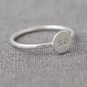 Può includere: Un anello color argento caratterizzato da una sottile fascia martellata e un piccolo disco circolare piatto sulla parte superiore. Il disco è stampato con le lettere S e O in un carattere semplice senza grazie. Il metallo ha una finitura opaca.