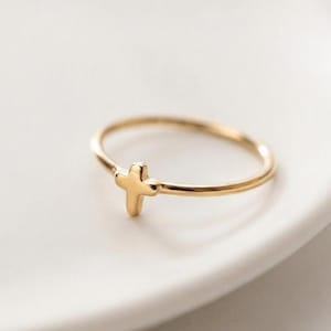 Peut inclure: Une fine bague en or ornée d'une petite croix centrée. L'anneau est lisse et circulaire, avec l'élément en forme de croix intégré directement dans la structure métallique, créant un bijou délicat et simple pour le doigt.