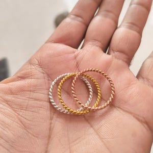 May include: A set of three thin, twisted metal rings in silver, gold, and rose gold tones. Each ring features a continuous rope-like texture throughout the band, creating a uniform, circular shape that is lightweight and simple in design.