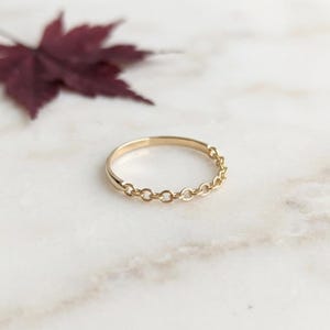 May include: A gold ring featuring a smooth, solid band on one side that transitions into a delicate chain-link design on the other side. The ring has a polished metallic finish and a circular shape, showcasing a refined and simple construction.
