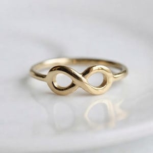 Op de afbeelding: Een gouden ring met een oneindigheidssymbool in het midden. De ringband is dun en rond en sluit aan de voorzijde aan op de horizontale acht-vorm. Het metaal heeft een gepolijst, reflecterend oppervlak met een warme gele kleur.