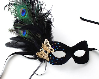 May include: Black masquerade mask with a peacock feather and gold butterfly embellishment. The mask is decorated with blue and gold rhinestones.