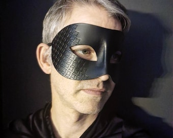 May include: A person wearing a black masquerade mask with a patterned design. The mask covers the person's eyes and part of their forehead. The person is wearing a black shirt.