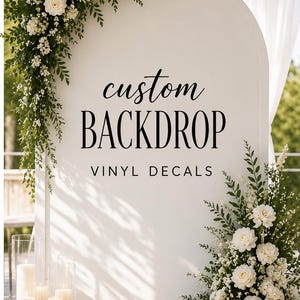 Puede incluir: Un panel de fondo arqueado blanco con el texto negro custom BACKDROP VINYL DECALS. La estructura está decorada con flores blancas y follaje verde en la parte superior izquierda e inferior derecha, junto a velas cilíndricas de cristal.