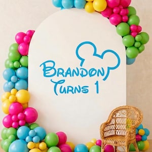 May include: A white arch-shaped sign with the text "Brandon Turns 1" in blue, surrounded by a colourful balloon garland. The balloons are blue, yellow, pink, and green. A small wicker chair sits in front of the sign, on a grey rug.