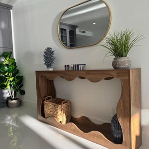 May include: A wooden console table with a wavy edge design, a gold-framed oval mirror, and decorative items. The table features a woven basket and a dark blue vase. A potted plant sits on the table, and another is on the floor.