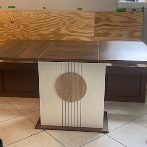 Puede incluir: Una mesa de comedor de madera con una tapa marrón oscuro y una base de madera clara. La base presenta un diseño circular y líneas verticales. La mesa está colocada contra una pared de color claro y una ventana.