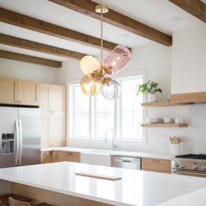 May include: A modern chandelier with a gold finish and multiple glass shades in various shapes and colors, including pink, amber, and clear. The fixture hangs from the ceiling in a kitchen with wooden beams and cabinetry.