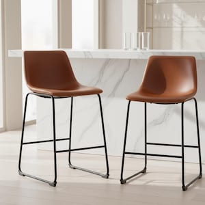 May include: Two brown leather bar stools with black metal frames. The stools have a curved seat and backrest, and a footrest. The stools are positioned in front of a white marble countertop.