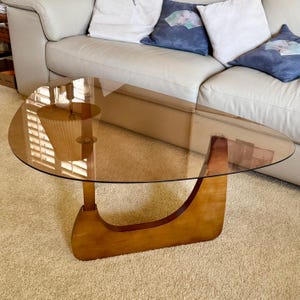 May include: A mid-century modern coffee table with a clear, oval-shaped glass top. The base is made of light-colored wood with a curved design. The table is set on a beige carpet in a living room setting.