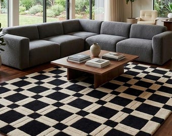 May include: A large rectangular area rug featuring a repeating geometric pattern of black and cream-colored squares and rectangles. The rug is positioned on a dark wood floor in front of a gray sectional sofa and a low wooden coffee table.