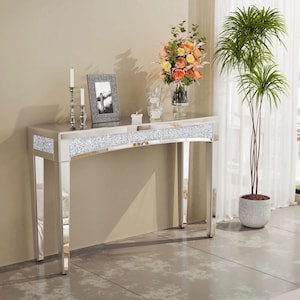Op de afbeelding: Een spiegelconsole met een gebogen voorkant en sprankelende kristallen accenten. De tafel heeft een reflecterend oppervlak en is versierd met kaarsen, een fotolijst en een vaas met bloemen. Rechts staat een potplant.