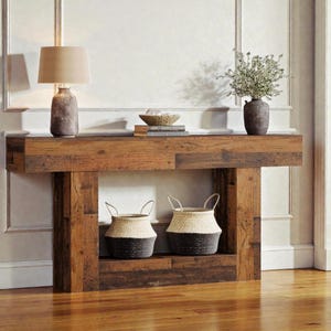 Peut inclure: Une console en bois rustique avec une lampe, des objets décoratifs et deux paniers tressés. La table a une finition en bois naturel et une étagère inférieure. Les paniers sont bicolores, avec un dessus clair et un fond foncé.