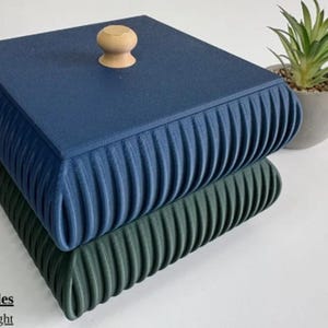 May include: Two square boxes, one navy blue and one dark green, stacked. The navy box has a wooden knob. The sides of the boxes have a ribbed design. A small potted plant is in the background. The boxes are likely for storage.