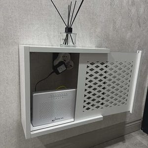 May include: A white wall-mounted cabinet with a decorative, diamond-patterned door. Inside, a modem and power adapter are visible. A glass diffuser with black reeds sits on top, adding a touch of decor to this functional storage solution.
