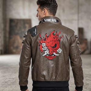 Puede incluir: Chaqueta de cuero marrón con un gráfico rojo y blanco en la espalda. La chaqueta tiene la palabra "SAMURAI" en el cuello y "CYBERPUNK 2077" en el hombro. Tiene puños de canalé negros y cintura negra.