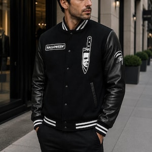 May include: A black varsity jacket with leather sleeves, featuring a white HALLOWEEN patch on the right chest and a white graphic of a knife with a face on the left chest. The jacket has a ribbed collar and cuffs with white stripes and snap buttons.
