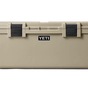 May include: A tan rectangular hard-sided cooler with a smooth finish. The lid features two black rubber latches on the front. A black rectangular badge with the white text YETI is centred on the front panel below the lid. Black handles are visible.
