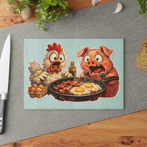 Puede incluir: Una tabla de cortar azul claro con una ilustración de dibujos animados de un pollo y un cerdo mirando una sartén con tocino y huevos. También hay una caja de huevos y una botella de aceite.