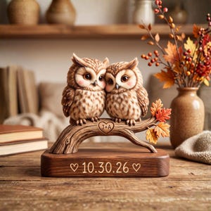 Può includere: Una statuetta decorativa con due gufi marroni appollaiati su un ramo con foglie autunnali. Il ramo è inciso con un cuore contenente le lettere J+A. La base in legno mostra la data 10.30.26 incisa, affiancata da due piccoli cuori.