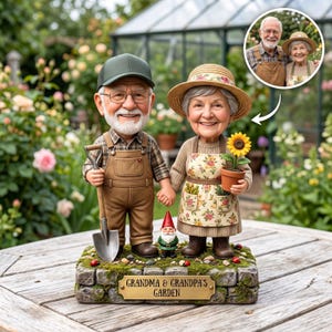 Puede incluir: Una figura personalizada de una pareja mayor sobre una base de piedra con una placa que dice GRANDMA & GRANDPA'S GARDEN. La figura masculina lleva una gorra verde, camisa a cuadros y peto marrón con una pala. La figura femenina lleva un sombrero de paja y un delantal floral con un girasol en maceta. Un pequeño gnomo de jardín se encuentra entre ellos.