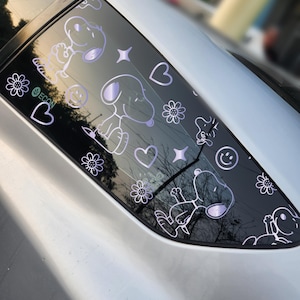 Puede incluir: Una calcomanía para ventana de coche con Snoopy y otros diseños de dibujos animados. La calcomanía negra tiene ilustraciones blancas y moradas de Snoopy, flores, corazones, estrellas y caras sonrientes. También es visible la palabra "eCON".