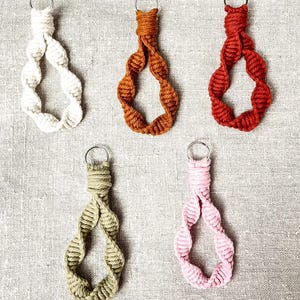 Könnte beinhalten: Fünf Makramee-Schlüsselanhänger in verschiedenen Farben: Weiß, Rost, Rot, Olivgrün und Rosa. Jeder Schlüsselanhänger hat ein gewebtes Design mit einem Metallring zum Befestigen von Schlüsseln. Die Schlüsselanhänger werden auf einem neutralen Leinenhintergrund präsentiert.