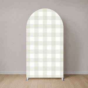 Puede incluir: Un soporte de fondo alto y arqueado con un patrón de cuadros vichy en color beige claro y blanco. La estructura tiene una parte superior redondeada y una base rectangular, apoyada por dos pequeños pies de metal en la parte inferior.