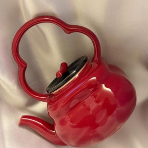 Könnte beinhalten: Glänzend rote Keramik-Teekanne mit herzförmigem Griff und schwarzem Deckel. Die Teekanne hat einen gebogenen Ausguss und einen kleinen roten Knopf auf dem Deckel. Schlichtes und elegantes Design, geeignet zum Servieren von Tee.