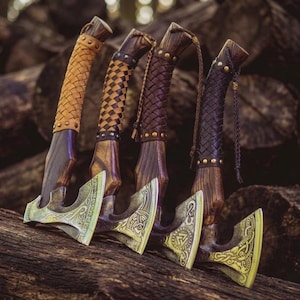 May include: Four handcrafted axes with wooden handles wrapped in leather. The axe heads have intricate designs and a metallic finish. The leather wraps are in various colours, including tan, brown, and black. The axes are displayed on a wooden surface.