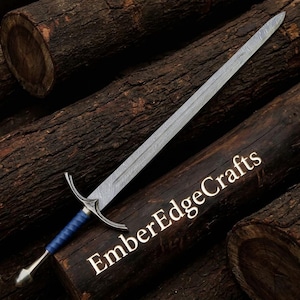 May include: A long, silver sword with a patterned blade and a blue handle. The sword has a curved crossguard and a rounded pommel. The text "EmberEdgeCrafts" is printed on the wooden surface.