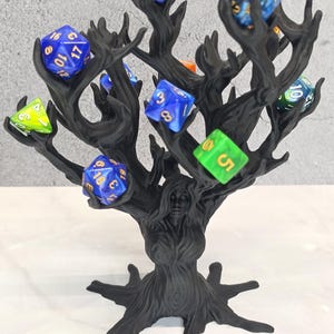 May include: A black, tree-shaped display stand with a central figure resembling a dryad or spirit, featuring multiple branches designed to hold polyhedral dice. Several multi-colored, translucent dice with gold-colored numbers are balanced on the branches.