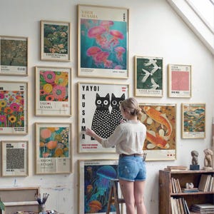 May include: A collection of framed art prints featuring various designs, including floral patterns, jellyfish, koi fish, and abstract art. The prints are displayed on a white wall, creating a gallery-like atmosphere. The artwork is by Yayoi Kusama.