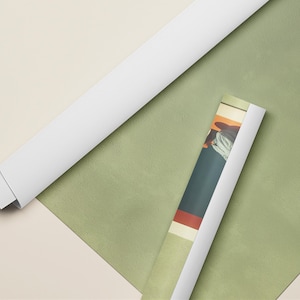 May include: Two rolled posters, one with a plain white back and one displaying a colorful graphic print featuring dark blue, red, and cream tones, resting on a flat, sage green surface with a partial view of a cream-colored frame edge.