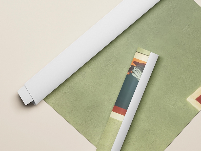 May include: Two rolled posters, one with a plain white back and one displaying a colorful graphic print featuring dark blue, red, and cream tones, resting on a flat, sage green surface with a partial view of a cream-colored frame edge.