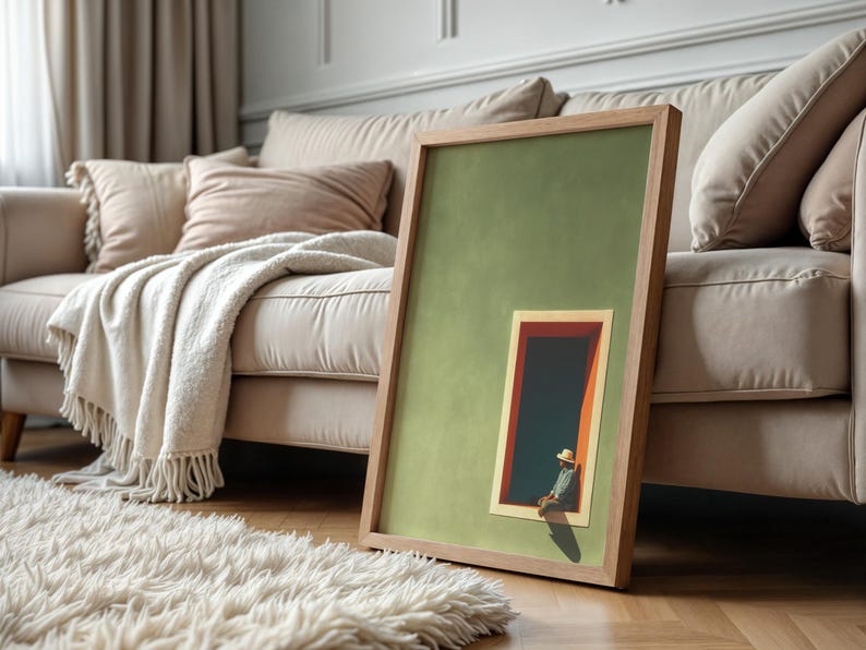 May include: A framed rectangular wall art piece featuring a solid sage green background with a smaller inset illustration of a person wearing a straw hat sitting in a dark doorway with orange trim, all enclosed in a light-toned wooden frame.