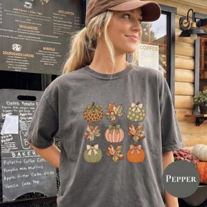 以下が含まれることがあります： チャコールグレーの半袖Tシャツ。リボン付きのカボチャ、柄入りの葉、無地のカボチャなど、ヒョウ柄、チェック柄、花柄を組み合わせた9つの秋をテーマにしたアイコンがグリッド状にプリントされています。
