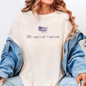 以下が含まれることがあります： 愛国的なデザインのクリーム色のTシャツ。赤、白、青でアメリカ合衆国の地図が描かれ、下に「250 years of freedom」の文字が書かれています。Tシャツはデニムジャケットと合わせて着用されています。