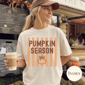 以下が含まれることがあります： 縦のオレンジ色のストライプと茶色の文字で「PUMPKIN SEASON」と書かれたグラフィックが特徴のアイボリーの半袖Tシャツ。文字の下には、茎にヒョウ柄のリボンが付いた薄いオレンジ色のカボチャのイラストが描かれています。