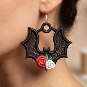 May include: A black bat-shaped earring featuring a textured wing pattern and a decorative cluster of one red rose, one white rose, and two green leaves at the bottom. The earring hangs from a silver-toned metal hook and has a circular cutout at the top.