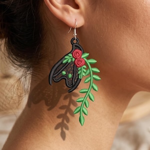 May include: A dangling earring featuring a black embroidered bat shape with two small red circular accents at the top, connected to a long, trailing green vine with multiple leaves, suspended from a silver-toned metal hook wire.