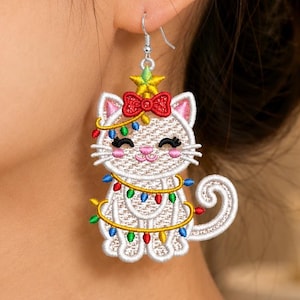 Puede incluir: Un pendiente bordado de gato que muestra un felino blanco con orejas y nariz rosadas, con un lazo rojo y una estrella amarilla en la cabeza. El gato está envuelto en una cadena de luces navideñas con bombillas rojas, azules, verdes y amarillas.