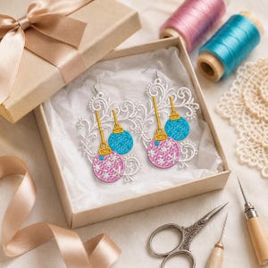 Puede incluir: Un par de pendientes bordados con filigrana blanca y formas de adornos navide&ntilde;os colgantes en rosa y azul. Los adornos tienen tapas doradas y patrones de copos de nieve blancos. Los pendientes se presentan en una caja de regalo.