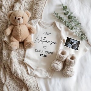 May include: A beige teddy bear, baby onesie, and knitted baby booties are arranged on a white surface. The onesie has the text "BABY Williamson DUE AUGUST 2026" in black script. A sonogram image and eucalyptus sprig are also present.